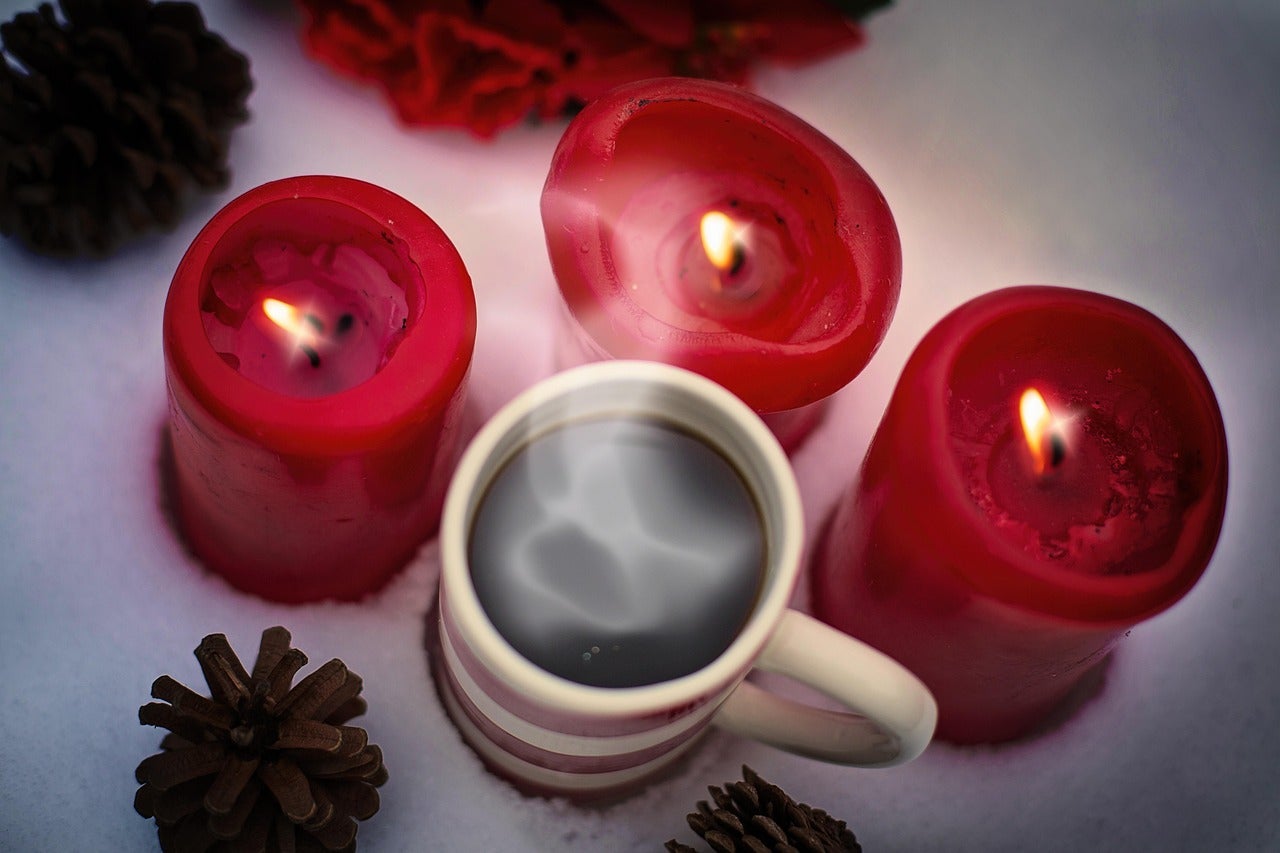 A mug of coffee nestled among three lit red candles and some pine cones.