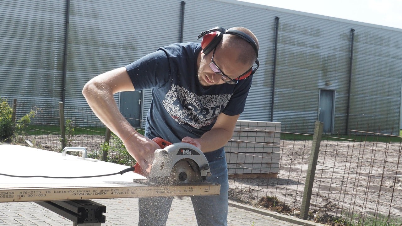 A person wearing safety glasses, ear muffs, and short sleeves using a circular saw to cut a door.